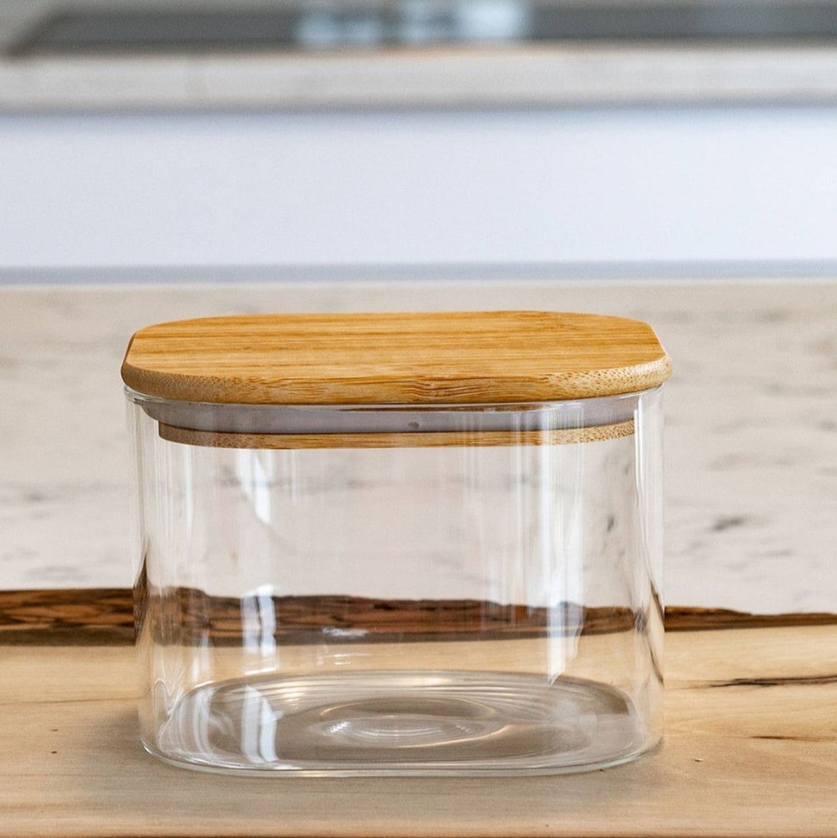 A clear glass storage jar with a light natural bamboo lid, set on a rustic wood slab with a blurred kitchen background.