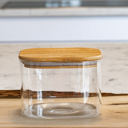 A clear glass storage jar with a light natural bamboo lid, set on a rustic wood slab with a blurred kitchen background.